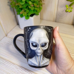 May include: Black ceramic mug with a hand-painted gray alien face. The alien has large black eyes and a serious expression.