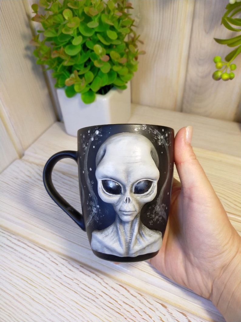 May include: Black ceramic mug with a hand-painted gray alien face. The alien has large black eyes and a serious expression.