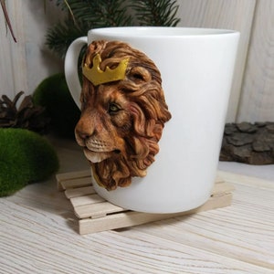 May include: White ceramic mug with a 3D sculpted lion head wearing a gold crown. The lion's mane is brown and the eyes are green.