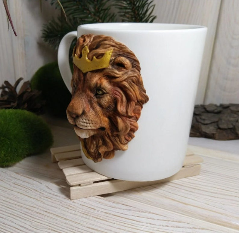 May include: White ceramic mug with a 3D sculpted lion head wearing a gold crown. The lion's mane is brown and the eyes are green.
