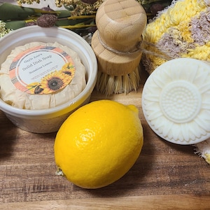 May include: A white ceramic dish filled with a round bar of solid dish soap with a sunflower design and the text " Gayle Ann's Solid Dish Soap Sunshine Lemon". Two yellow lemons, a white round soap with a floral design, and a wooden brush with natural bristles are also pictured.
