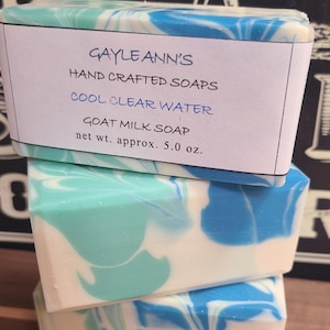 May include: Three bars of handmade goat milk soap, stacked on top of each other. The top bar has a label that reads "Gayle Ann's Hand Crafted Soaps Cool Clear Water Goat Milk Soap net wt. approx. 5.0 oz.". The soap is a swirl of white, blue, and green.