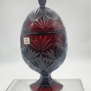 以下が含まれることがあります： ルビーレッドの蓋付きガラス瓶。扇のような模様が施されています。瓶は卵形で、六角形の台座に置かれています。側面のステッカーには「MADE IN FRANCE」と書かれています。