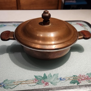 May include: A round copper cooking pot with a matching lid, featuring two dark brown wooden side handles and a matching dark brown wooden knob on the center of the lid. The pot has a metallic copper finish with visible signs of wear and patina.