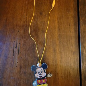 May include: A yellow string necklace with a Mickey Mouse charm. The charm is made of plastic and features Mickey Mouse in his signature red shorts and yellow shoes.