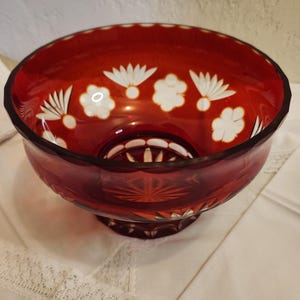 May include: A ruby red glass bowl with a decorative floral pattern. The bowl has a round shape with a wide rim and a pedestal base. White floral designs are etched around the rim. The bowl is sitting on a white surface.