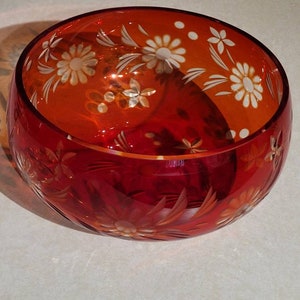May include: A round, red glass bowl with etched floral designs. The bowl has a wide opening and a thick base. The etched flowers are white and are arranged around the rim and sides of the bowl. The bowl is sitting on a white surface.