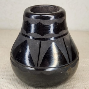 May include: A small, black ceramic vase with a rounded base and a wide opening. The vase features etched geometric patterns, including triangles and scalloped lines, adding a decorative touch to the design. The vase is likely handmade.
