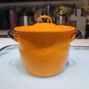 May include: Orange enamel pot with black handles and a lid. The pot is sitting on a white surface.