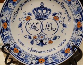 May include: A white ceramic plate with a blue and orange floral design. The plate features the text "Willem-Alexander ∞ Maxima Zorreguieta" and "2 februari 2002" along with a crown and the initials "W" and "M" intertwined.