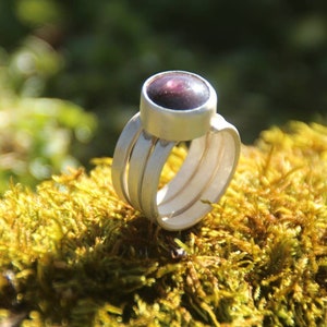 Könnte beinhalten: Ein silberner Ring mit einem dunkelvioletten Edelstein, der in einer Zargenfassung gefasst ist. Der Ring hat drei Bänder, die übereinander gestapelt sind.