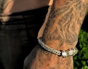 May include: A silver braided bracelet with a clasp fastening. The bracelet is worn on a person's wrist with a tattoo of a lion's face.
