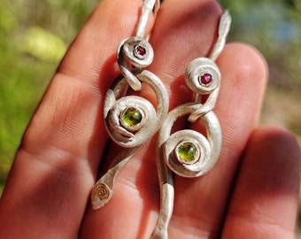 May include: A pair of silver earrings with a twisted design. Each earring features a green and red gemstone.