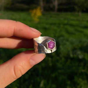 May include: A silver ring with a hammered texture and a hexagonal, magenta-colored gemstone. The ring is held up against a blurred green background, showcasing the jewelry's design and color.