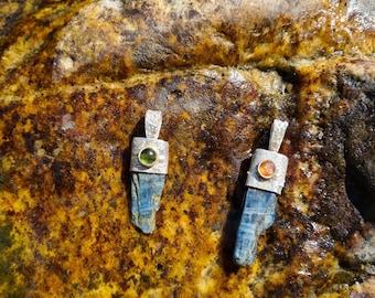 May include: Two pendant necklaces with blue kyanite crystals and small, round gemstones. The pendants have silver-colored metal settings. One gemstone is green, and the other is orange. The pendants are displayed on a textured, yellow and brown surface.