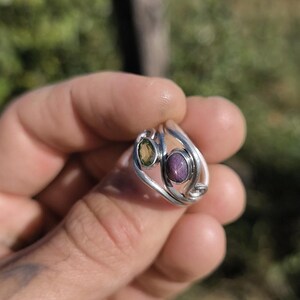 May include: A silver ring featuring a bypass design with two oval-shaped stones. One stone is a translucent olive green, and the other is a deep purple with a visible star-like inclusion. The metal band has a smooth, polished, and reflective finish.