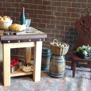 May include: A miniature wooden table with a chequered top, a bowl of fruit, a loaf of bread, a bottle of wine, and two glasses. The table has a shelf underneath with a small box of red apples and a jar labelled "Honey". There are two barrels and a chair with a carved back in the background.