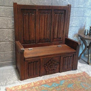 Può includere: Una panca in miniatura in legno con schienale alto e sedile incernierato. La panca è di colore marrone scuro e presenta dettagli intagliati sul davanti. La scena comprende un tavolino, un candelabro e un tappeto.