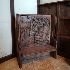 May include: A wooden bench with a carved scene of a cabin in the woods. The bench is dark brown and has a simple design.