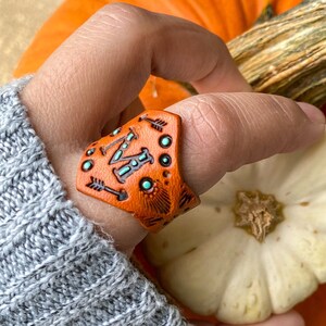 以下が含まれることがあります： オレンジ色の背景とターコイズのアクセントが付いた革のリング。リングには、スタイリッシュな文字「M」と矢印が描かれています。