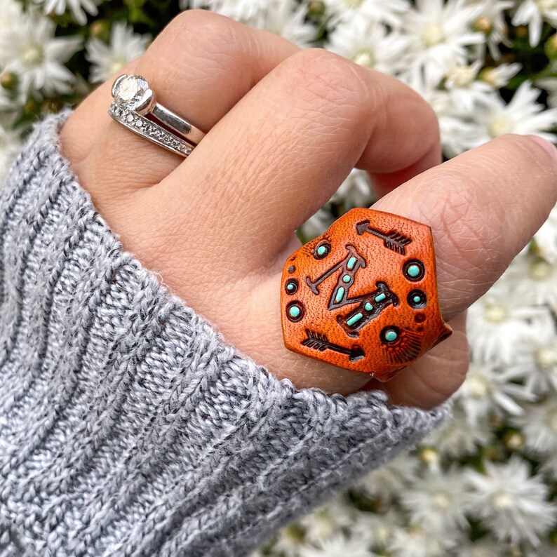 以下が含まれることがあります： ターコイズのアクセントと矢印のデザインが施された茶色の革のリング。リングは指につけられています。