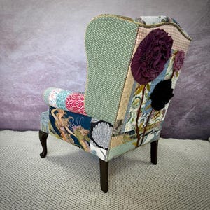 May include: A patchwork wingback chair with a variety of patterned fabrics in shades of green, blue, pink, and beige. The chair features dark wooden legs and is adorned with large, fabric flower embellishments in purple and black.