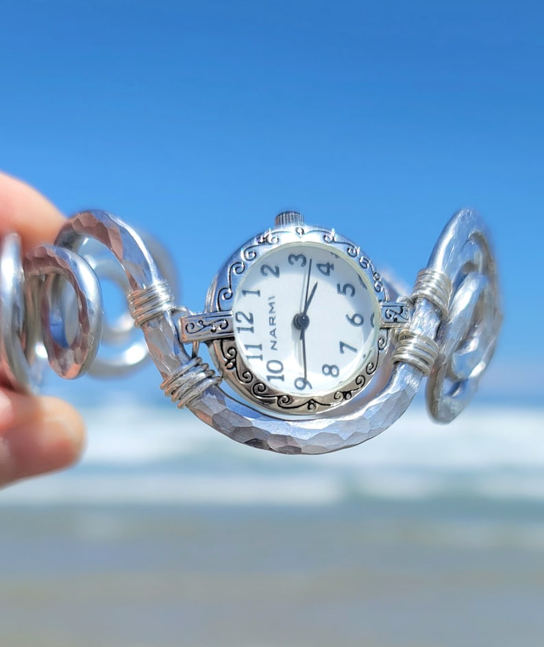 K&ouml;nnte beinhalten: Eine silberfarbene Uhr mit wei&szlig;em Zifferblatt und schwarzen Ziffern. Die Uhr hat eine dekorative L&uuml;nette und ist an einem geh&auml;mmerten Metallarmband befestigt. Das Zifferblatt zeigt "NARMI". Der Hintergrund ist eine verschwommene Ansicht von Meer und Himmel.