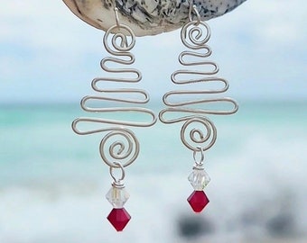 May include: A pair of silver wire earrings with a unique, abstract design. Each earring features a swirling top and a zig-zag pattern, with a clear crystal and a red, diamond-shaped bead dangling at the bottom. The earrings are displayed against a blurred ocean backdrop.