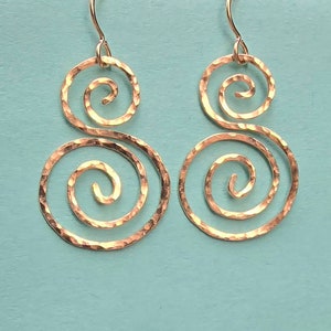 May include: A pair of hammered copper spiral earrings. Each earring features two connected spirals, with a textured surface and a hook for pierced ears. The earrings are displayed against a light blue background.