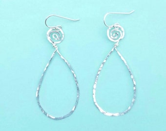 May include: A pair of silver-colored, hammered-texture teardrop earrings. Each earring features a spiral design at the top, connected to a teardrop shape. The earrings hang from a simple hook. The background is a light blue.