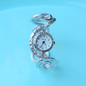K&ouml;nnte beinhalten: Silberfarbene Geneva-Uhr mit wei&szlig;em Zifferblatt und schwarzen Ziffern. Die Uhr hat ein einzigartiges, geh&auml;mmertes Metallband, das sich spiralf&ouml;rmig um das Handgelenk wickelt. Das Zifferblatt hat eine dekorative silberne L&uuml;nette.