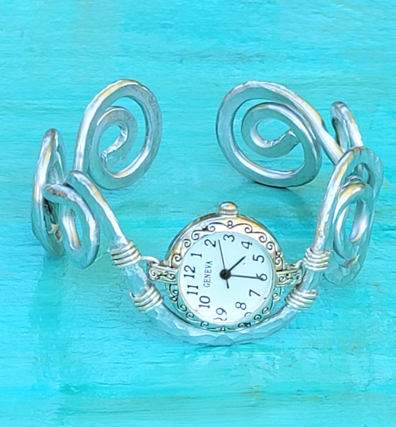 K&ouml;nnte beinhalten: Silberfarbene Armbanduhr mit wei&szlig;em Zifferblatt und schwarzen Zeigern. Das Zifferblatt hat schwarze Ziffern und den Schriftzug "GENEVA". Das Armband hat eine geh&auml;mmerten Textur und dekorative Wirbel.