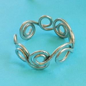 May include: A silver-colored metal bracelet with a series of spiral designs. The bracelet is circular and appears to be handmade. The background is a solid light blue.