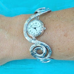 K&ouml;nnte beinhalten: Eine silberfarbene Geneva-Uhr mit wei&szlig;em Zifferblatt und schwarzen Ziffern. Die Uhr ist an einem einzigartigen, geh&auml;mmerten Metallarmband mit Spiraldesign befestigt. Das Armband ist etwa 17.78 cm lang.