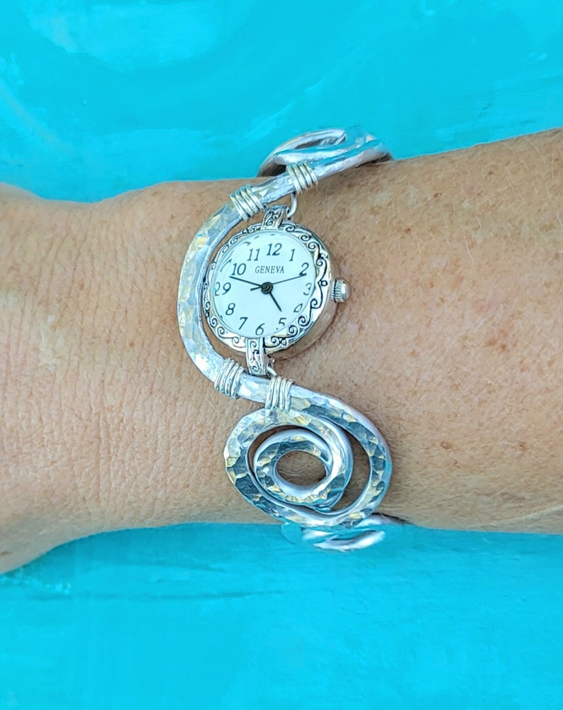 K&ouml;nnte beinhalten: Eine silberfarbene Geneva-Uhr mit wei&szlig;em Zifferblatt und schwarzen Ziffern. Die Uhr ist an einem einzigartigen, geh&auml;mmerten Metallarmband mit Spiraldesign befestigt. Das Armband ist etwa 17.78 cm lang.