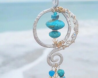 May include: A silver-toned pendant featuring a hammered circular frame with gold-toned wire accents. The centre holds two turquoise-coloured beads, while the bottom spiral section is adorned with dangling turquoise, clear, and white beads on a cord.