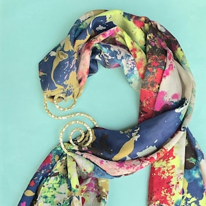 May include: A colorful scarf with a gold-toned metal scarf ring. The scarf features a vibrant abstract design with splashes of blue, yellow, red, and green. The scarf ring has a decorative, swirling design.