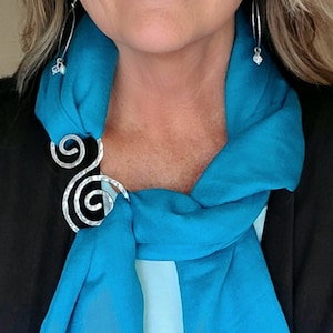 May include: A silver scarf clip with a double spiral design secures a bright turquoise scarf. The scarf is draped over a black top, and silver hoop earrings with small charms are visible.