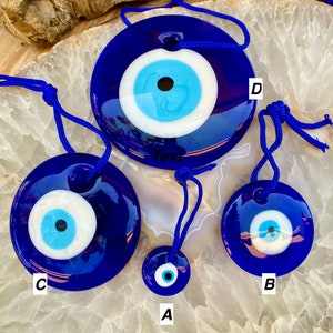 May include: Four blue glass evil eye charms with white and blue eyes and a black pupil. The charms are hanging from blue cords. The charms are arranged in a circle on a white and brown surface. The largest charm is labeled "D", the next largest is labeled "C", the next is labeled "B", and the smallest is labeled "A".