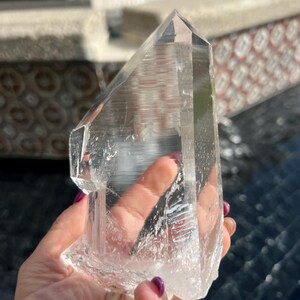 May include: A large, clear quartz crystal with a pointed tip and a rough, textured base. The crystal is held up against a blurred outdoor background, showcasing its natural clarity and crystalline structure. This item is a natural gemstone.