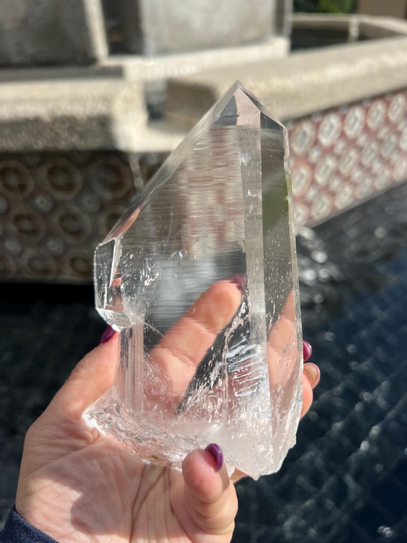May include: A large, clear quartz crystal with a pointed tip and a rough, textured base. The crystal is held up against a blurred outdoor background, showcasing its natural clarity and crystalline structure. This item is a natural gemstone.