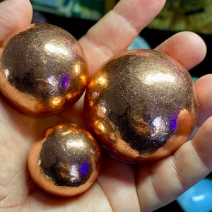 May include: Three copper spheres of varying sizes, with a shiny, metallic finish. The spheres are smooth and have a warm, reddish-brown color.