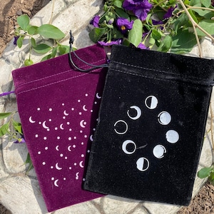May include: Two velvet pouches with drawstring closures. One pouch is black with a white moon phase design. The other pouch is burgundy with a white crescent moon and star pattern.