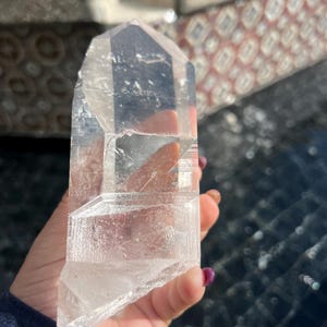 May include: A clear quartz crystal point, held in a hand. The crystal is transparent with visible internal structures and facets. The crystal is approximately 5 inches in length. The background is blurred, with hints of a patterned surface.