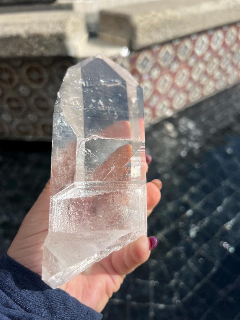 May include: A clear quartz crystal point, held in a hand. The crystal is transparent with visible internal structures and facets. The crystal is approximately 5 inches in length. The background is blurred, with hints of a patterned surface.