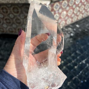 May include: A large, clear quartz crystal with a pointed top and a rough, textured base. The crystal is transparent, allowing light to pass through, and is held up in front of a background.
