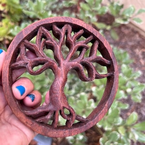 May include: A round wooden carving of a tree of life with intricate details. The carving is dark brown and has a natural wood finish.