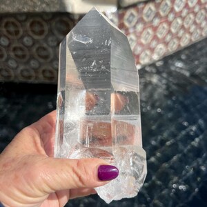 May include: A clear quartz crystal point with a faceted top and a rough, natural base. The crystal is held in a hand, showcasing its transparency and the light reflecting through its structure. The crystal is a natural, unpolished gemstone.