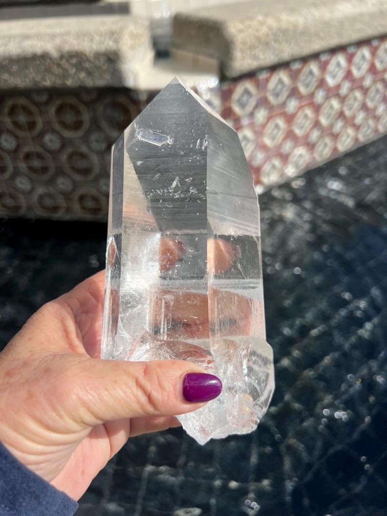May include: A clear quartz crystal point with a faceted top and a rough, natural base. The crystal is held in a hand, showcasing its transparency and the light reflecting through its structure. The crystal is a natural, unpolished gemstone.