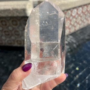 May include: A clear quartz crystal point with a faceted, geometric shape. The crystal is held in a hand with purple nail polish. The crystal is approximately 6 inches tall.